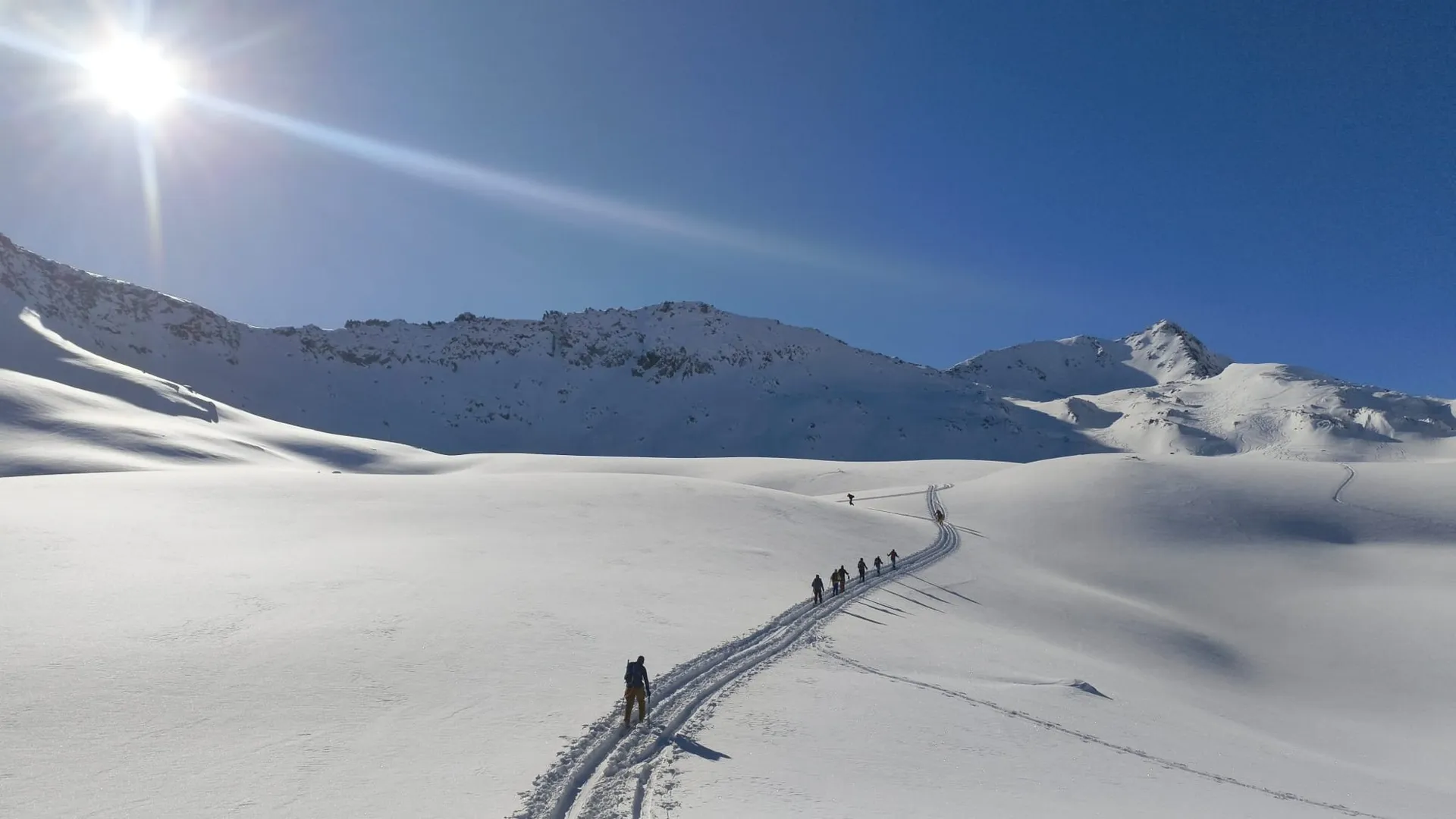 A line of ski tourers skinning up a wide snowfield under a blue sky