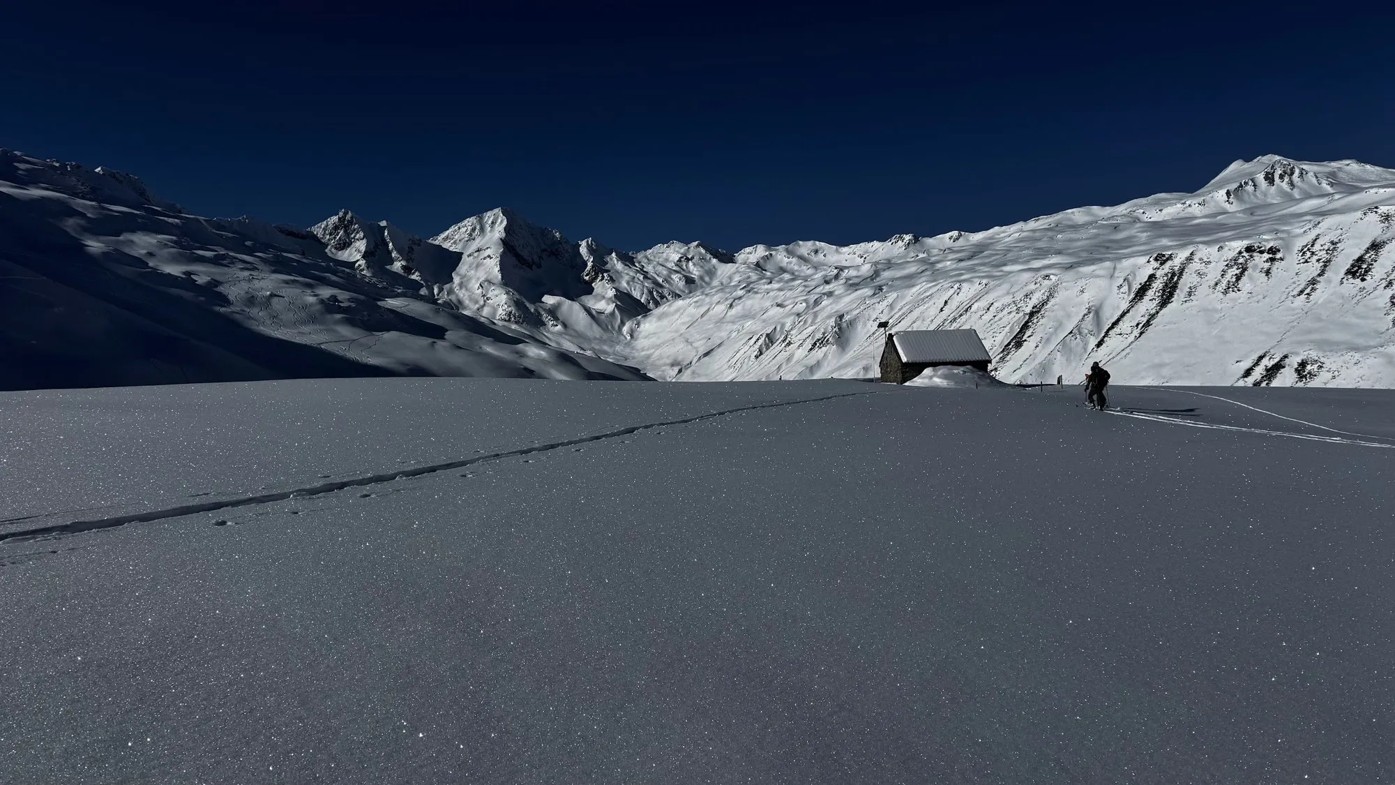A ski tourer passing a small snow-covered stone hut, with white peaks behind