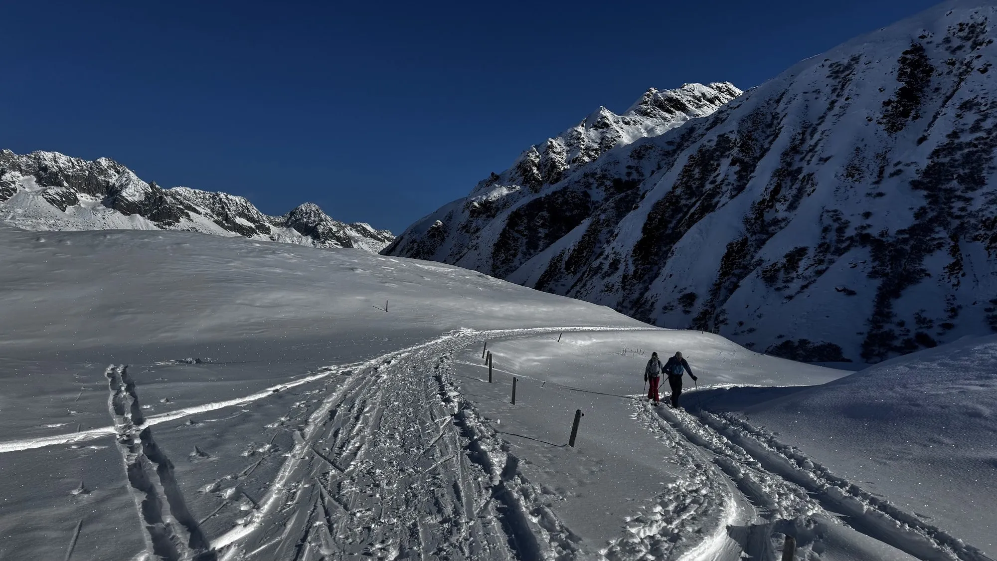 Two ski tourers on a snowy track above Realp, with jagged peaks behind